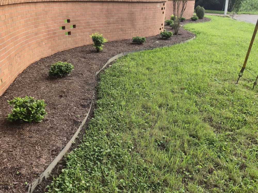 Curved mulch bed with small shrubs and edging along a brick wall, with green lawn beside it