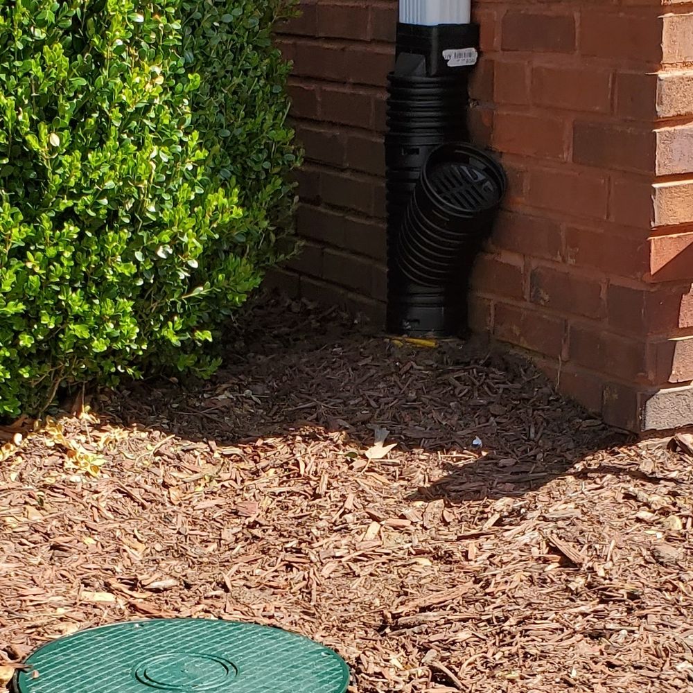 Black corrugated pipe connected to a downspout at the corner of a brick wall, running into a fresh mulch bed, with a round green lid nearby