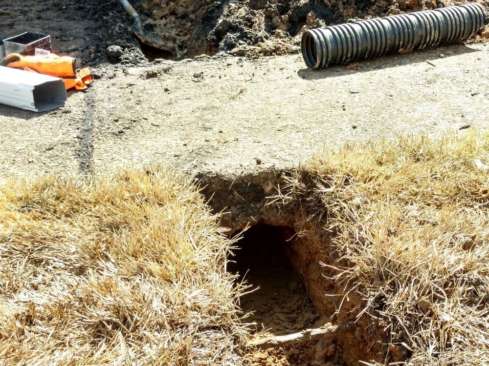Trench dug through dry grass along the edge of a concrete slab, with black corrugated drain pipe laid out nearby