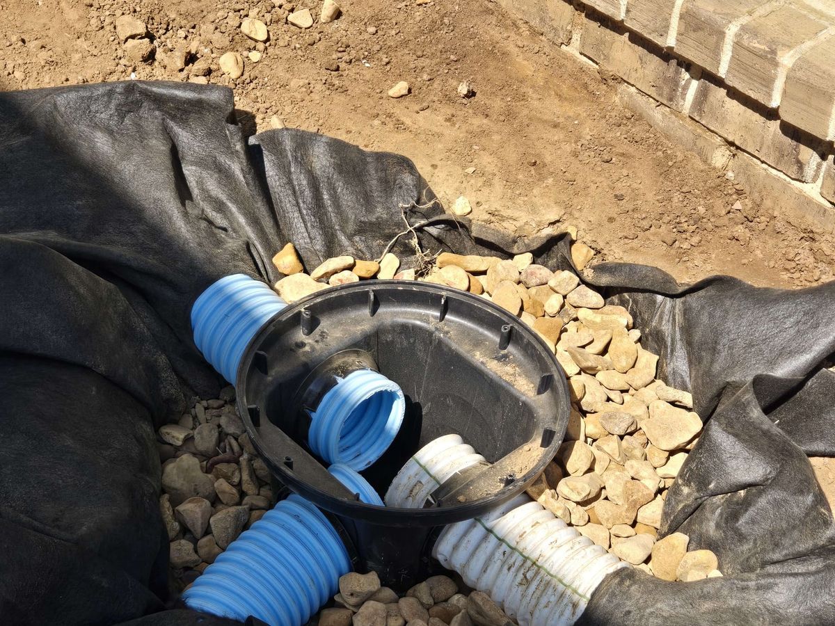 Black drainage basin connected to blue and white corrugated pipes, set in gravel on landscape fabric next to a brick edge
