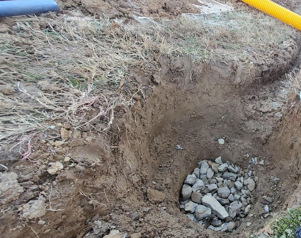 Dug hole with gravel at the bottom and a trench leading toward yellow corrugated drain pipe in a lawn