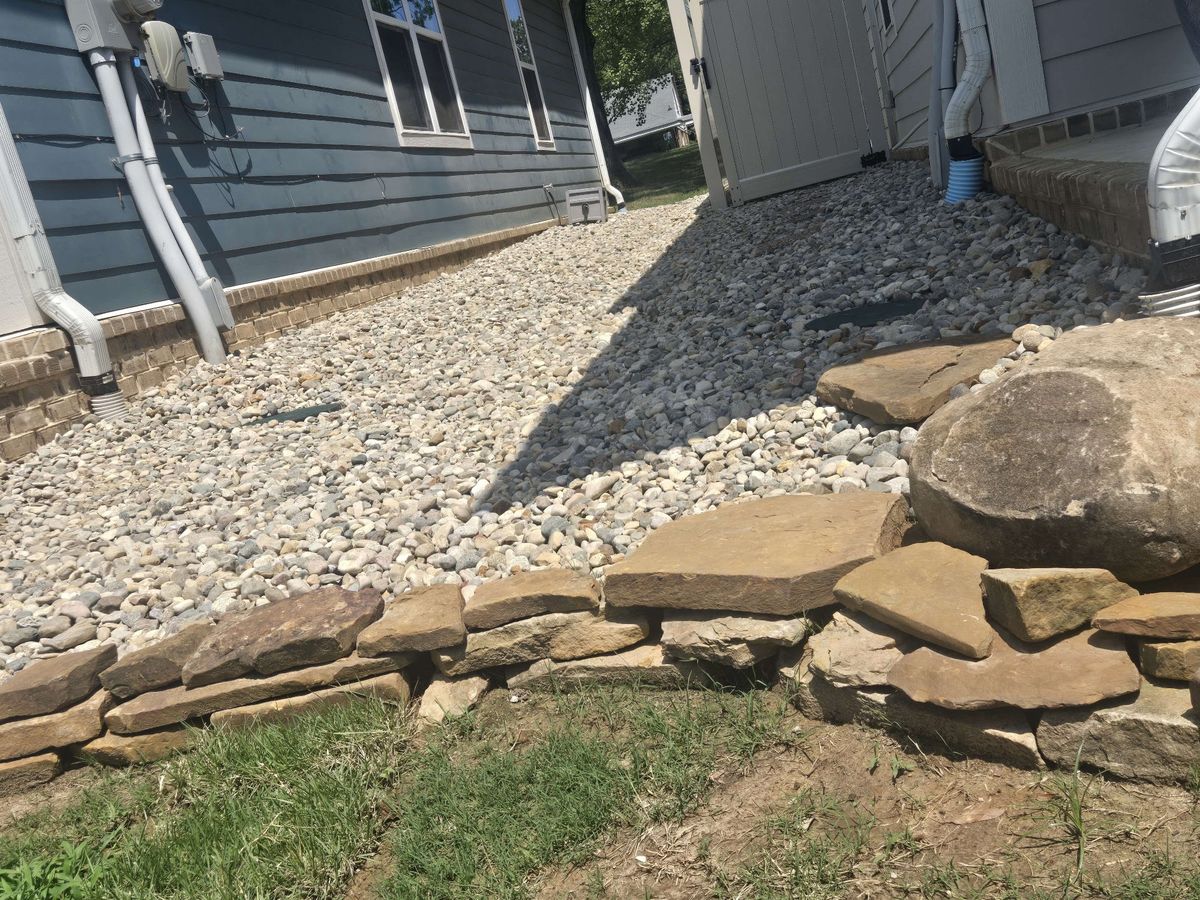 Gravel bed along gray house siding with a downspout, edged with stacked flagstone and boulders
